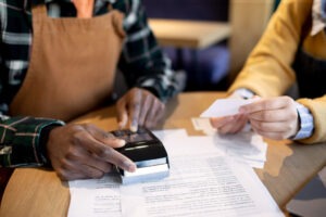 Duas pessoas interagindo em ambiente de comércio, com maquinha de cartão e tipos de notas fiscais sobre a mesa e na mão de uma das pessoas