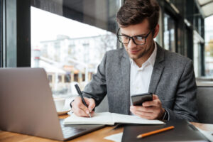 Empresário com terno e óculos de grau usando celular em frente a um notebook e fazendo anotações e um caderno, simbolizando formas de solicitar inscrição estadual